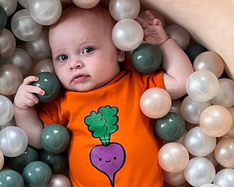 Baby in an orange onesie with a purple and green vegetable design, surrounded by colorful balls.
